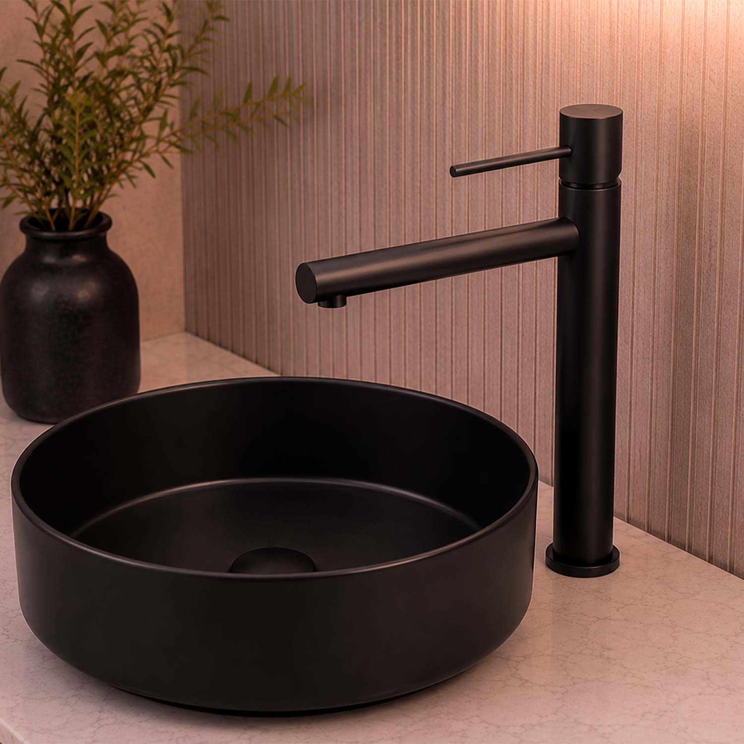 Matte black vessel sink with a matching single-lever faucet on a light stone countertop, with a dark vase of greenery in the background.