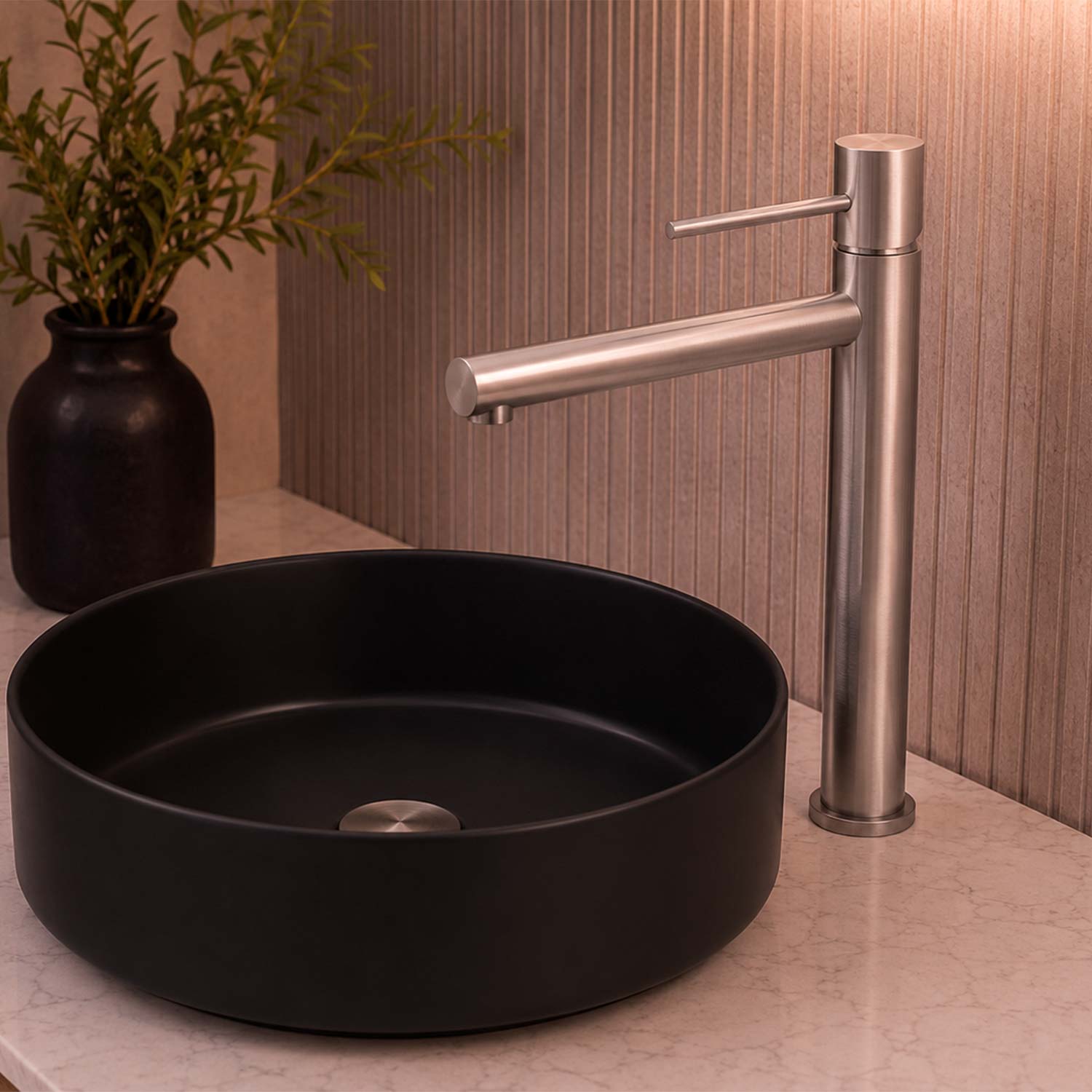 A matte black round vessel sink paired with a modern tall faucet in brushed silver, set on a light stone countertop against a textured beige wall with a small potted plant in the background.