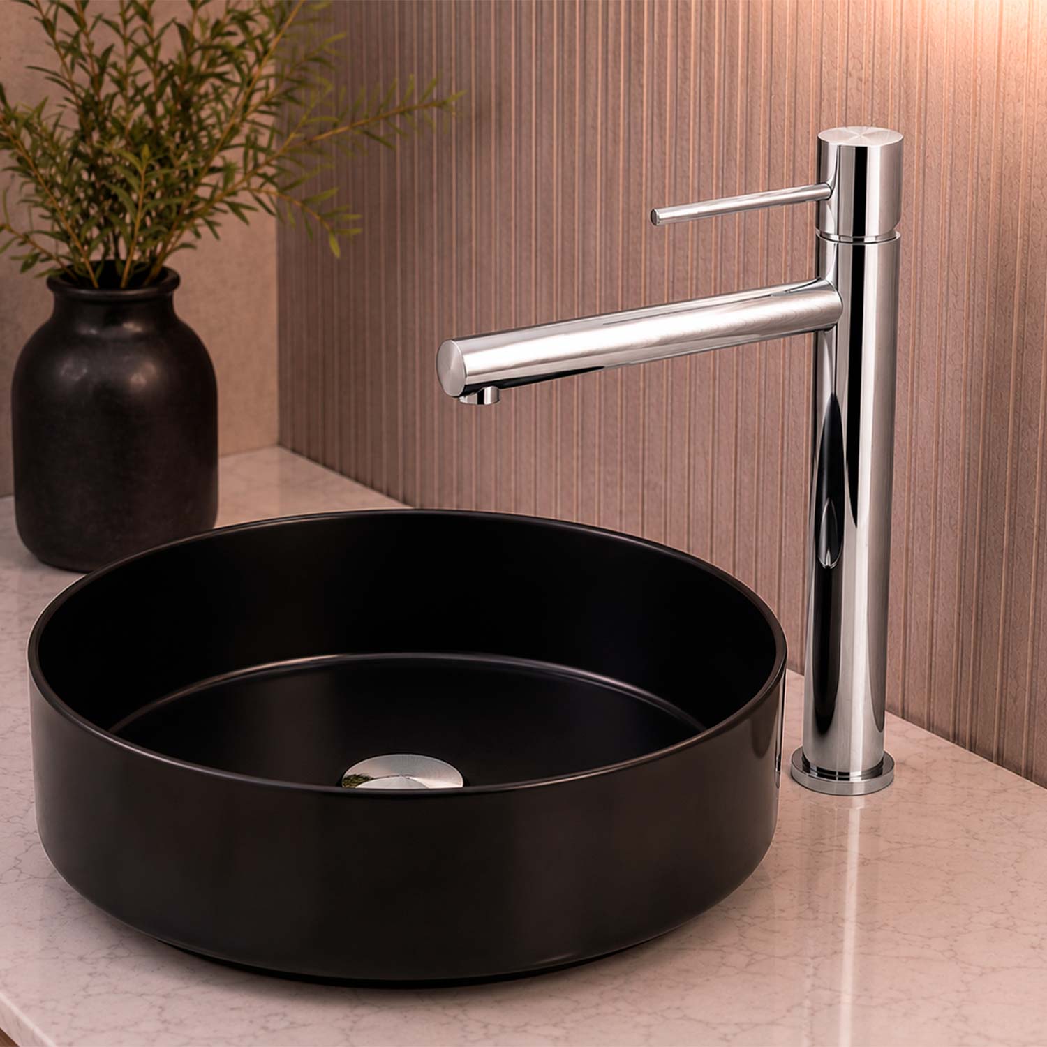 A modern bathroom sink setup with a round matte black vessel basin and a tall polished chrome faucet, shown on a light stone countertop with a brown textured wall and a dark vase of greenery in the background.