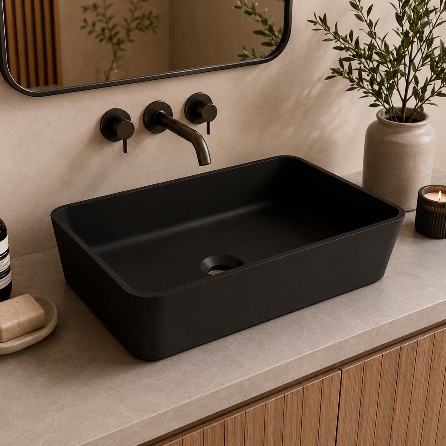 A matte black rectangular vessel sink installed on a light stone countertop in a bathroom setting, with a matching dark faucet, beige wall, mirror, plant in a vase, and a small candle.